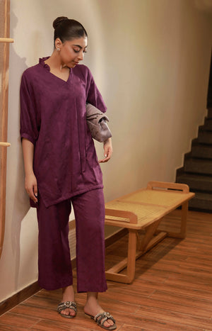Full shot of the plum purple tunic and trouser set styled with a shoulder handbag.

