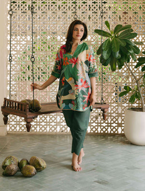 Woman in a colorful floral dress standing next to a wooden swing with plants in a decorative setting.

